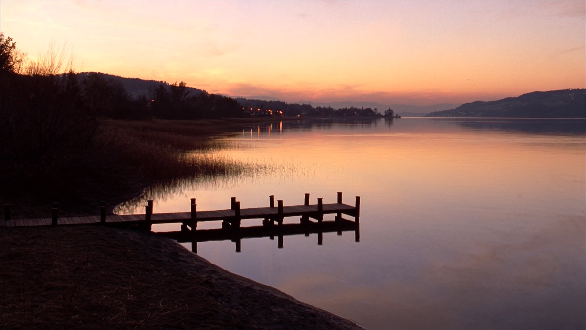 Steg am Greifensee in der Abenddämmerung: Schilf im Vordergrund, spiegelglattes Wasser im letzten Abendrot, Uferlichter am gegenüberliegenden Seeufer — der See, an dem Uster liegt.
