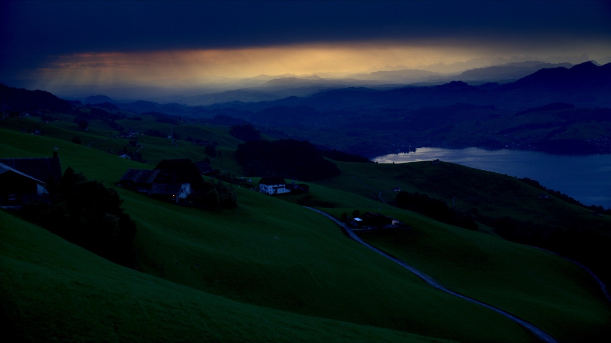 Hügellandschaft im Zürcher Oberland in der Abenddämmerung: grüne Kuppen, verstreute Bauernhöfe und ein schimmernder See im Tal unter dramatischem Himmel.